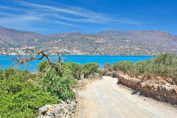 Naklejka premium A dirt road in Kalydon (Spinalonga) peninsula near the Elounda town in Crete, Greece. Beautiful blue sky with white clouds in the background.