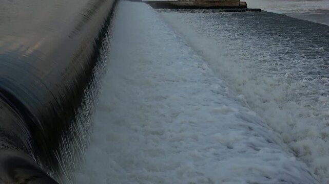 Close-up of clean river water sliding smoothly over the curved surface of an inflated black rubber dam before bursting into active white bubbles.
