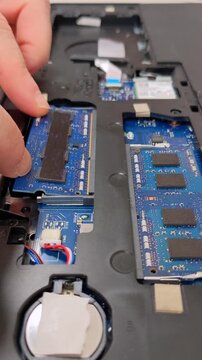 A technician's fingers position a SO-DIMM module over an exposed circuit board, with chips, connectors, and the CMOS battery visible