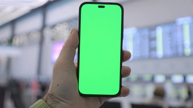 Hand holds phone with green screen in public space. Background blurred with bright lights and indistinct figures. Suitable for app demos, digital content, or marketing visuals