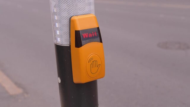 Close-up of a person's hand pressing a yellow pedestrian crossing button on a traffic light pole, activating the signal to cross the street safely while waiting for cars to stop