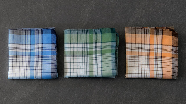 Composition of three classic men's cotton handkerchiefs in blue, green, and orange plaid patterns on a dark background.