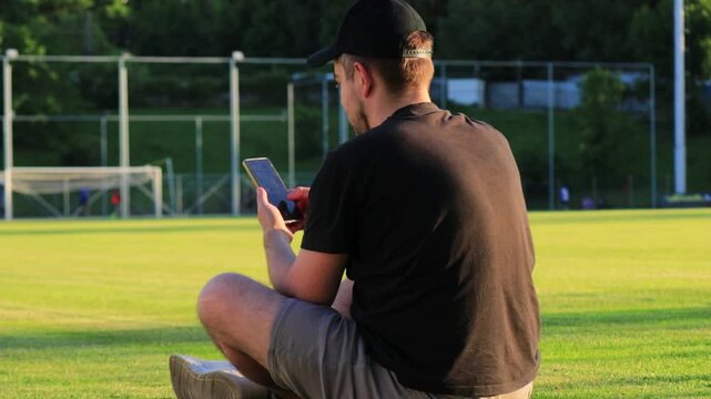 White man sitting on grass at a football stadium browsing smartphone while other players play football in the background at sunset. Concept of phone addiction, social isolation, social anxiety, sports
