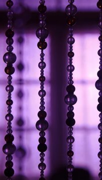 Vertical video: Swaying beaded curtain turning at window by breeze, catching amber above dark shape