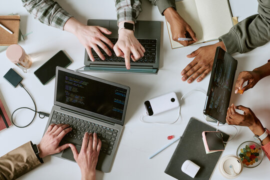 Overhead shot of diverse young adult team coding on laptops and tablet, sharing notes during software project collaboration. Ideal for tech teamwork, startup, agile development marketing