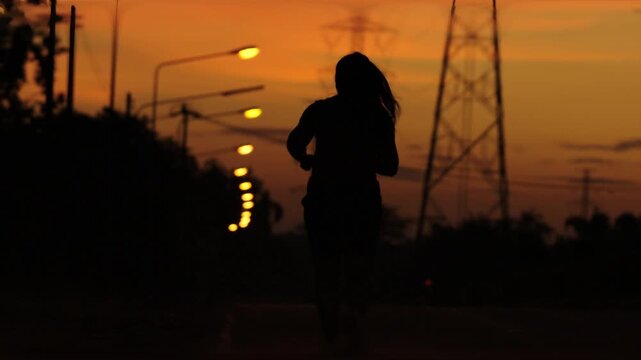 Cinematic slow motion silhouette of a woman running on an empty street at dusk under warm sunset light, creating a moody urban atmosphere with street lamps and power lines in the background