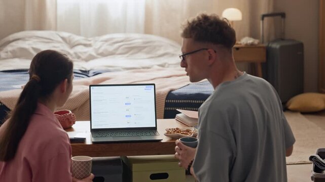 Back view of young adult Caucasian couple sitting in bedroom, using laptop to plan travel and book flights. Footage is perfect for online booking services and tourism marketing campaigns