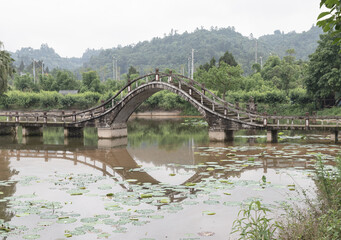 Obraz premium In Dazu Lotus Manor, a high-arched, traditional stone footbridge gracefully spans a calm body of water. Its distinctive, steeply curved silhouette casts a mirrored reflection onto the surface below, w