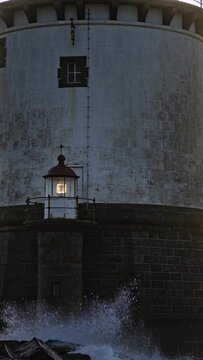 Vertical video: Facing incoming swells, lanterned tower at rocky jetty taking waves, creating spray