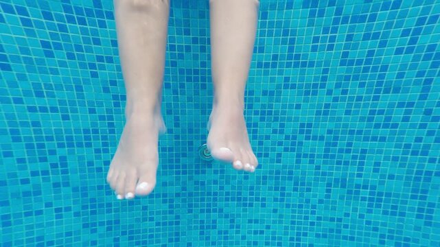Female legs with white pedicure dangling in the clear blue water of a swimming pool