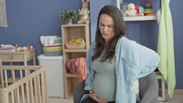 Pregnant woman holds belly with one hand and presses lower back for backache in nursery; maternal discomfort.