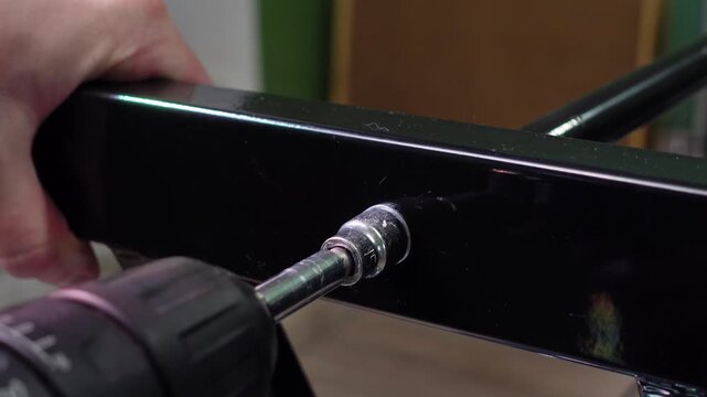 A person using an electric cordless drill with a socket wrench attachment to unscrew a bolt on a black metal folding chair frame