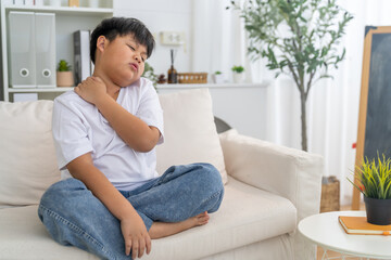 Fototapeta premium Sad asian boy suffering from neck pain and shoulder ache sitting on sofa at home, Unhappy child massaging tense muscles feeling physical discomfort, Healthcare, poor posture, physical injury concepts