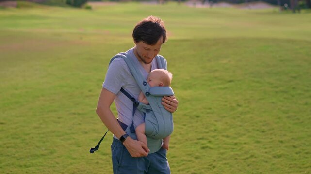 Father walking with baby in ergonomic carrier across green field. Slow motion.