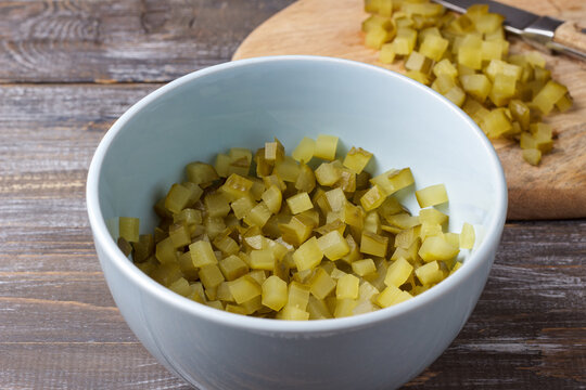 Finely diced pickles in a bowl with chopped cucumber pieces on a cutting board, prepared as an ingredient for cooking on a rustic wooden surface
