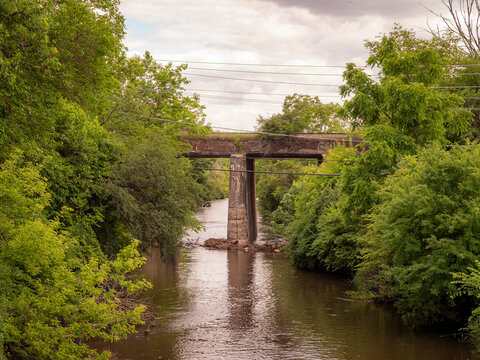 An old concrete railroad bridge spanning over a river surrounded by lush green summer trees and foliage under a cloudy sky