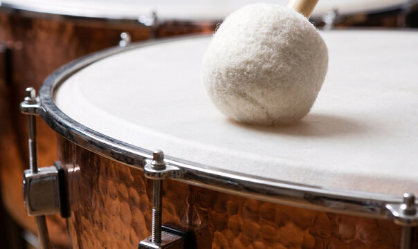 soft, padded felt mallet striking the stretched white skin of a large copper timpani drum