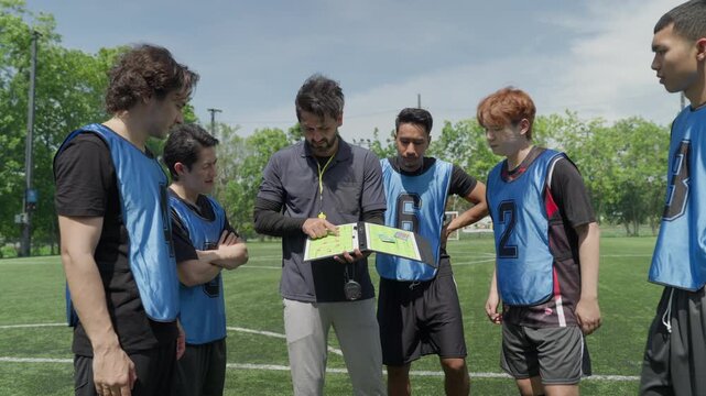 Professional soccer coach explaining tactic game plan on clipboard to team of young players. Diverse football athletes listening to trainer strategy during training match outdoor