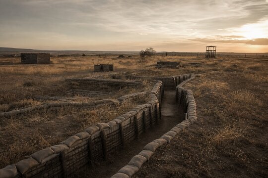 Deserted military training ground with sandbag fortifications and overgrown grass under golden hour light.