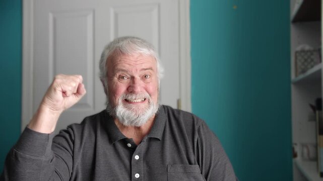 An elderly man makes several facial expressions and gestures while sitting indoors. He raises his fist and smiles at the camera. The setting is simple with a light background.