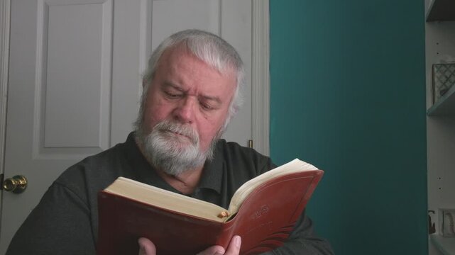 An older man sits in a room while reading a book. He looks focused as he turns the pages. The light comes in through a nearby window. He enjoys his time reading at home after lunch.