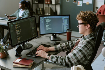 Young adult Caucasian man coding at dual monitors in office, developing software and testing interface. Ideal for IT recruitment, programming education, tech teamwork campaigns © AnnaStills