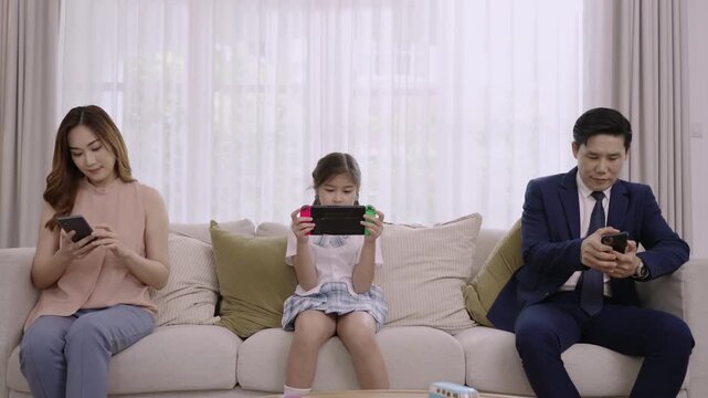 Asian family devices addicted to and overuse social media, so they are not paying attention to each other father and mother playing smartphones while daughter play game console on sofa in living room.