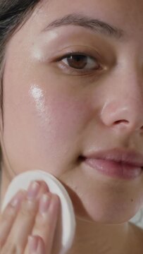 Vertical video: Bringing cotton pad, Asian woman toning cheeks at sink with right hand, in towel