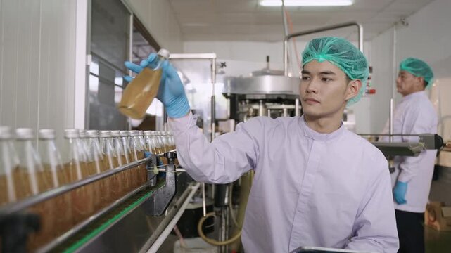 Asian quality control inspector in a uniform food and drink factory holding a tablet inspecting fruit juice bottles while moving on conveyor belt in beverage production line at manufacturing industry.
