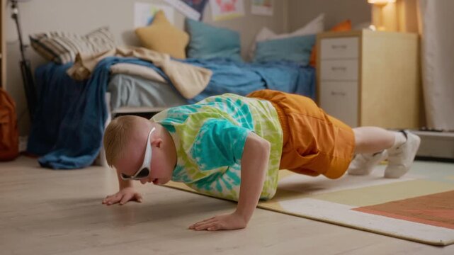 Caucasian boy with Down syndrome wearing sunglasses doing pushups on floor in bedroom. Footage ideal for campaigns promoting inclusivity, adaptive fitness, and diversity in youth sports