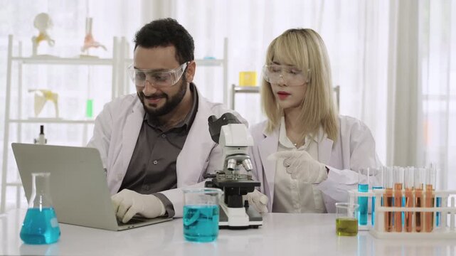 Two happy professional scientists and researchers in lab coats using a microscope and a laptop while conducting chemistry experiments with liquid in test tubes and beakers in the science laboratory. 