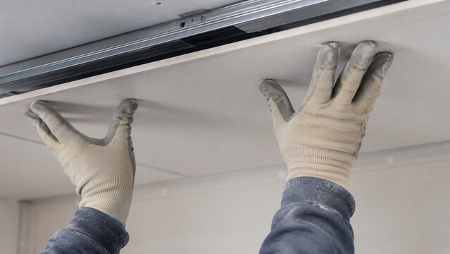 Worker wearing gloves installs a drywall panel on a ceiling, demonstrating construction or renovation work.