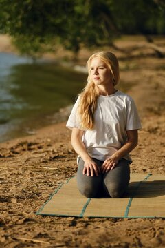 A woman sits on a mat by the riverside, engaged in a moment of mindfulness. She has long blonde hair and wears a simple white t-shirt and gray leggings. The warm sun illuminates the peaceful setting