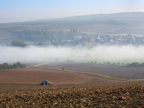 View of the village of Rhoden in autumn, fog sweeping through the valley, a tractor ploughs a field, Osterwieck, Harz, Harz foothills, Saxony-Anhalt, Germany