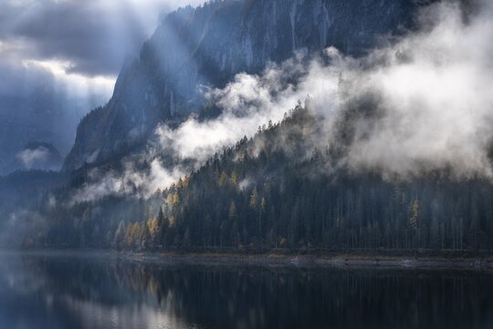 The Vordere Gosausee in autumn with a view of the Gosaukamm. Rays of sunshine shine through the clouds. Forest shrouded in clouds. Vorderer Gosausee, Gosau, Gosau valley, Salzkammergut, Upper Austria, Austria