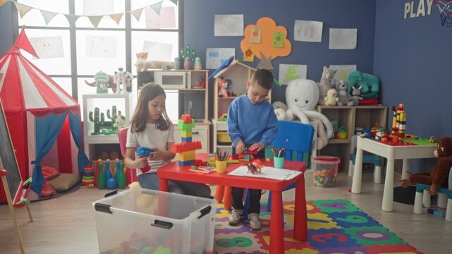 Girl and boy stacking colorful plastic blocks into a tall tower on a red table while crayons and a clear toy bin sit nearby in a playroom studio; creative joy.