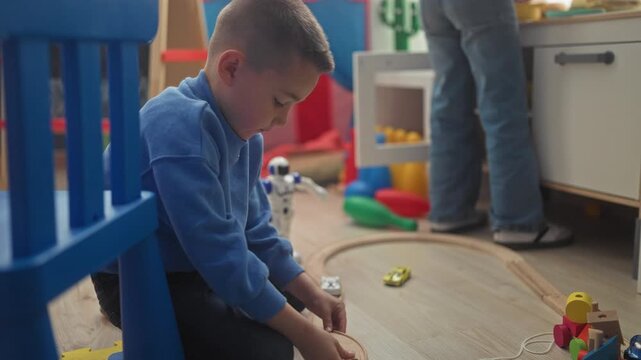 Boy kneeling on floor, hands assembling a wooden train track and pushing a toy car in a playroom of a building; quiet concentration.