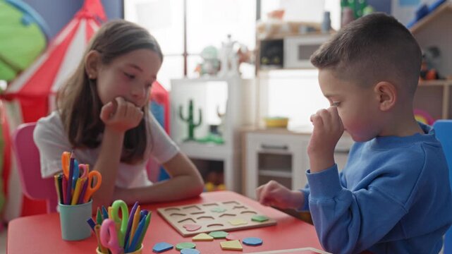Girl and boy at table play with wooden shape puzzle, boy places geometric pieces while girl rests chin on hand in a preschool building; quiet concentration.