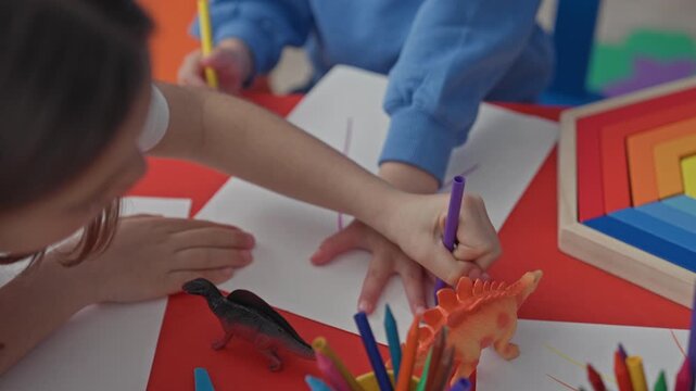 Girl traces another child's hand with a purple marker at a studio table with toy dinosaurs, colored pencils and paper visible; playful creativity.