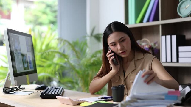 Millennial asian businesswoman talking on smartphone while managing a large pile of documents and analyzing financial data on a computer screen in a modern bright office