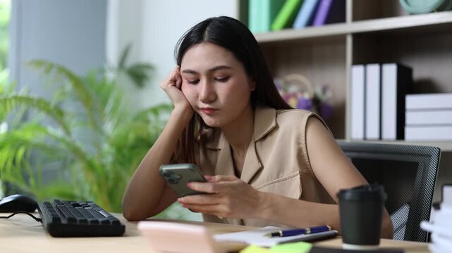 Bored young asian businesswoman using a smartphone at her desk, procrastinating at work and feeling unmotivated while browsing social media instead of working on a project