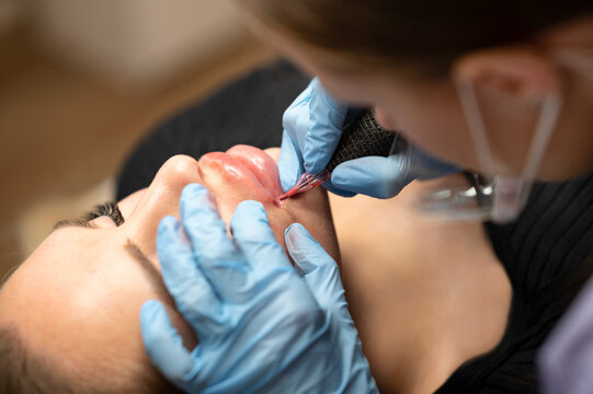 Close up of beautician in blue gloves applying permanent makeup on female lips. Cosmetic lip tattoo procedure in beauty salon. Professional artist working with tattoo machine