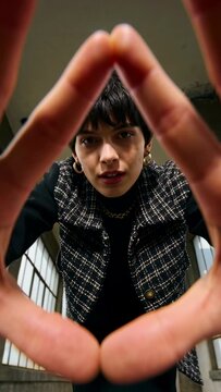 Vertical video: Leaning woman reacting to low camera, framing face with hands in loft, showing vest