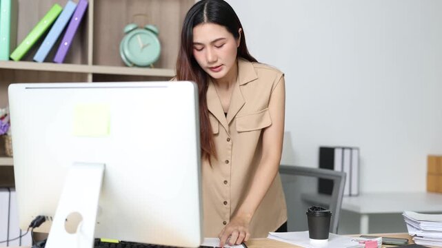 Young Asian businesswoman entrepreneur typing on a computer keyboard and working with documents on a desk, smiling at the camera in a modern bright corporate office