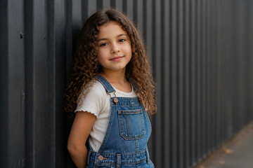 Fototapeta premium A young girl with curly brown hair wearing denim overalls stands against a dark corrugated metal fence.