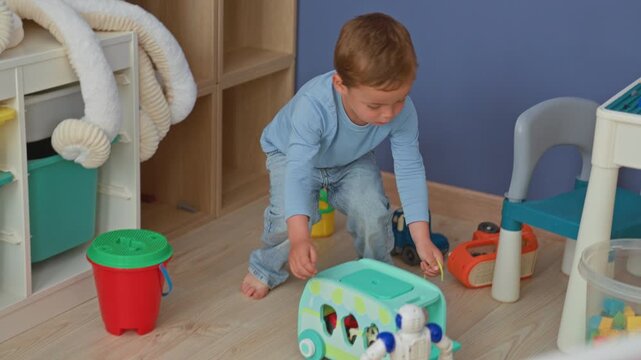 Boy pulls a string on a toy bus and bends to pick up a robot on the floor in a building; focused curiosity.