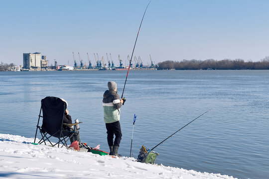 Winter fishing on snowy riverbank with angler and industrial harbor background