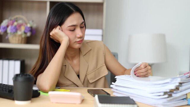 Bored and tired office worker feeling sleepy and unmotivated while sorting through a large pile of documents and paperwork at her messy desk in the workplace