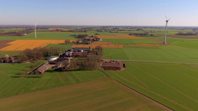 Aerial view of a traditional countryside farm and wind turbines in green fields. Drone landscape of agricultural farmlands, country houses, and wind generators under a clear blue sky, panorama.