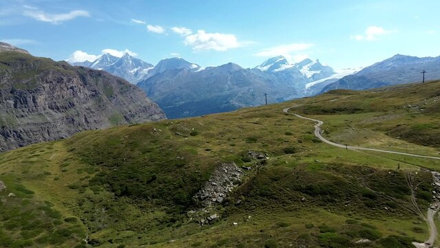 Aerial view of the Matterhorn Glacier landscape with rugged mountain peaks, green alpine meadows, and a winding dirt path under a clear blue sky in Zermatt, Valais, Switzerland.